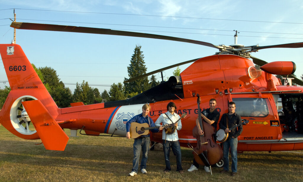 The Bluegrass band, Northern Departure, is pictured here in Brier, Washington, standing in front of a U.S. Coast Guard helicopter holding their instruments; an acoustic  guitar/vox, banjo/vox, bass/vox, and mandolin/vox.