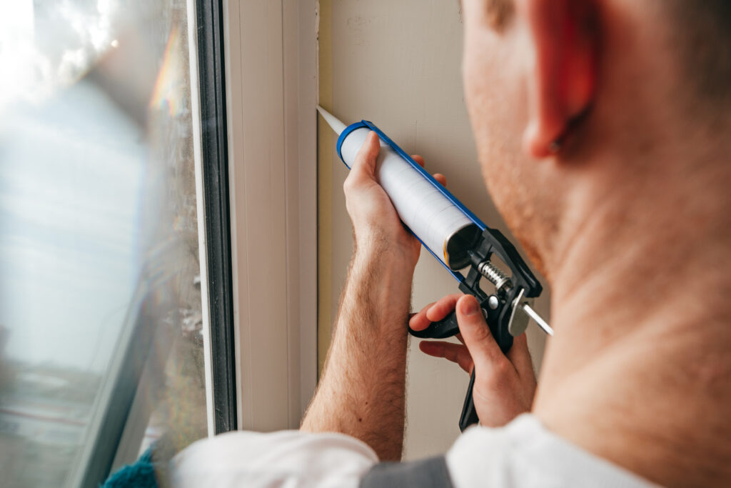 Man sealing cracks between window and trim, reducing air leakage around doors and windows is a good step to take in your home whether you are getting solar panels or not and can drastically improve energy efficiency.